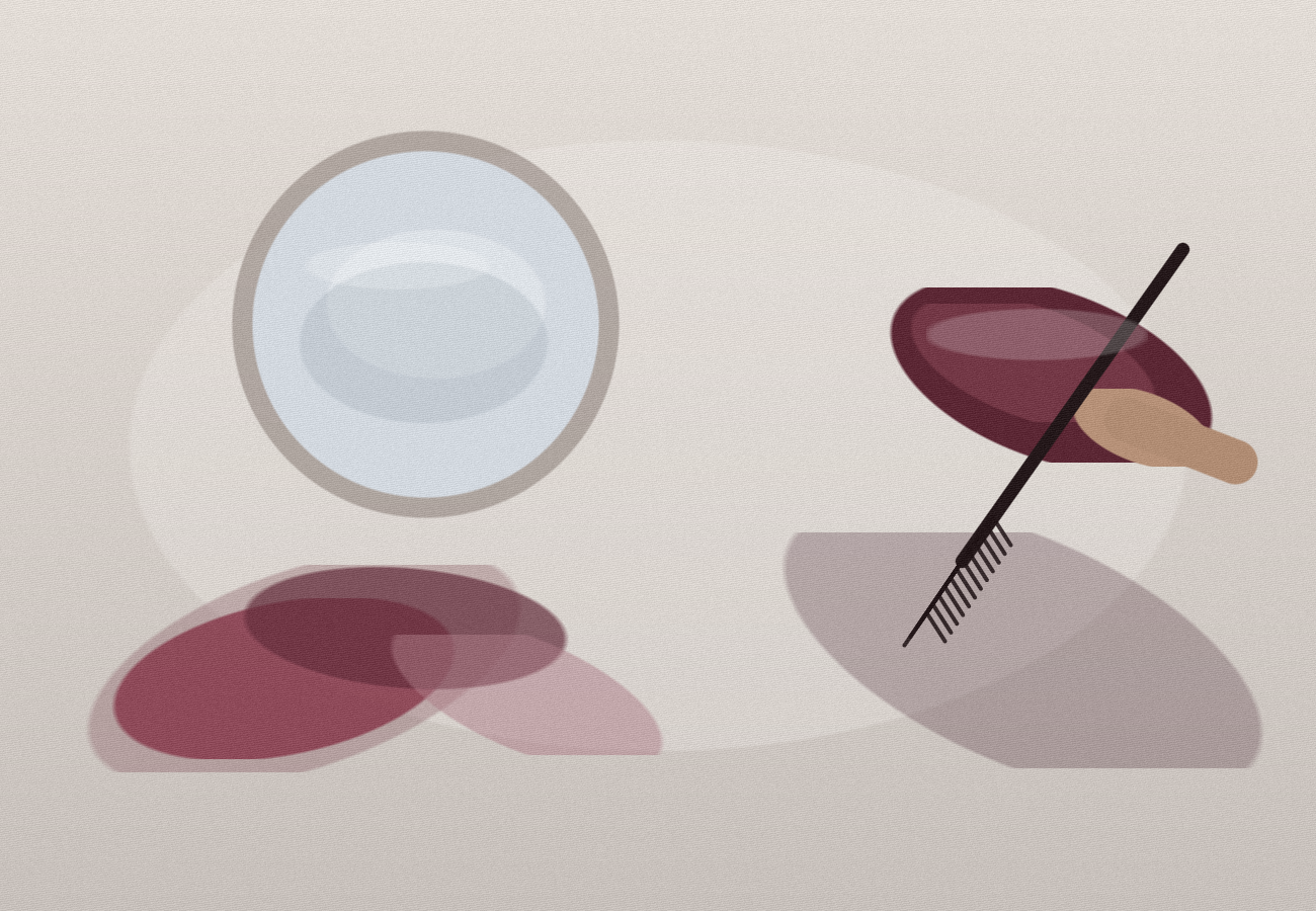 A burgundy mascara tube and wand rest beside an open compact mirror on a stone vanity with berry-toned fabric.