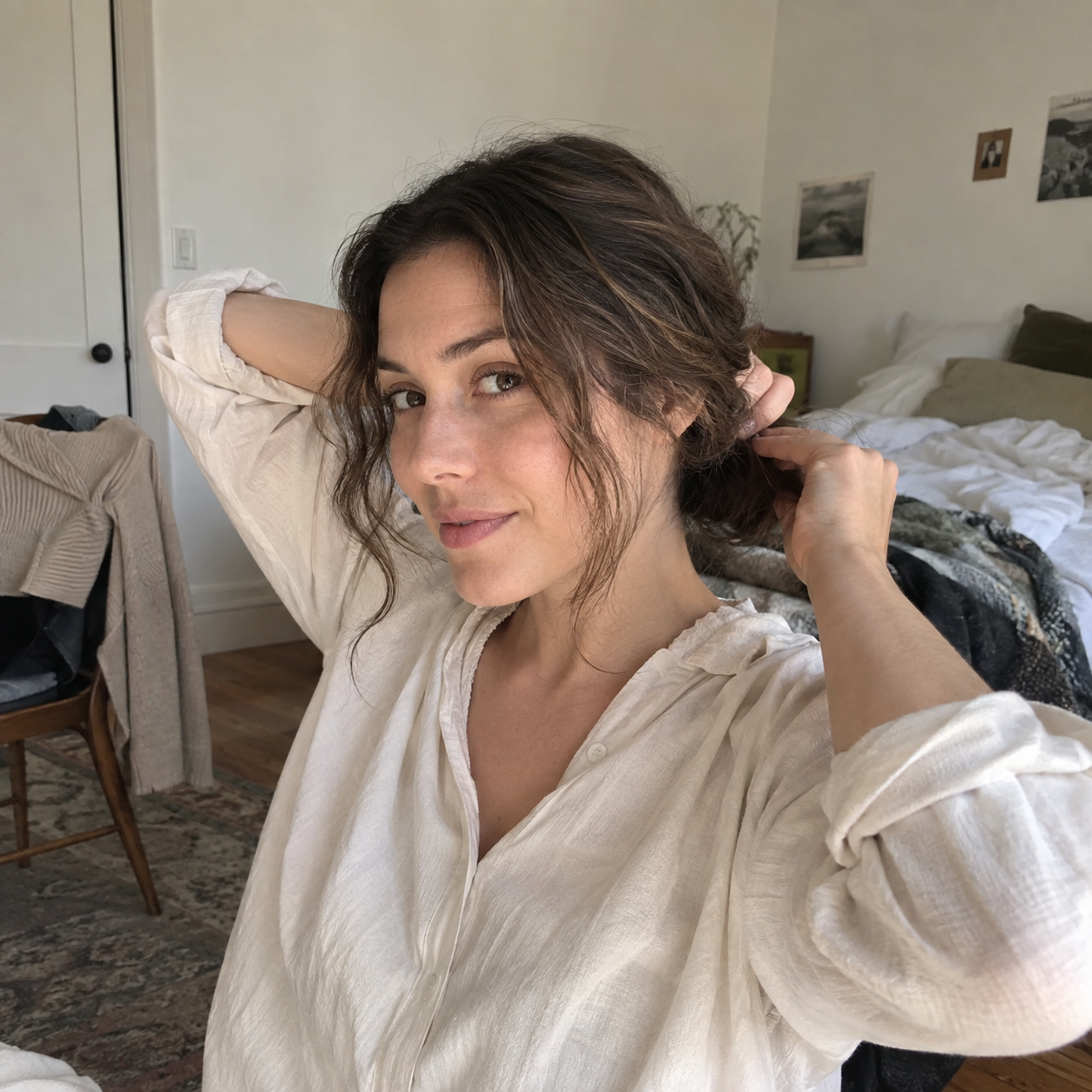 Woman in a cream shirt gently gathering her brown hair at the nape in a lived-in bedroom.