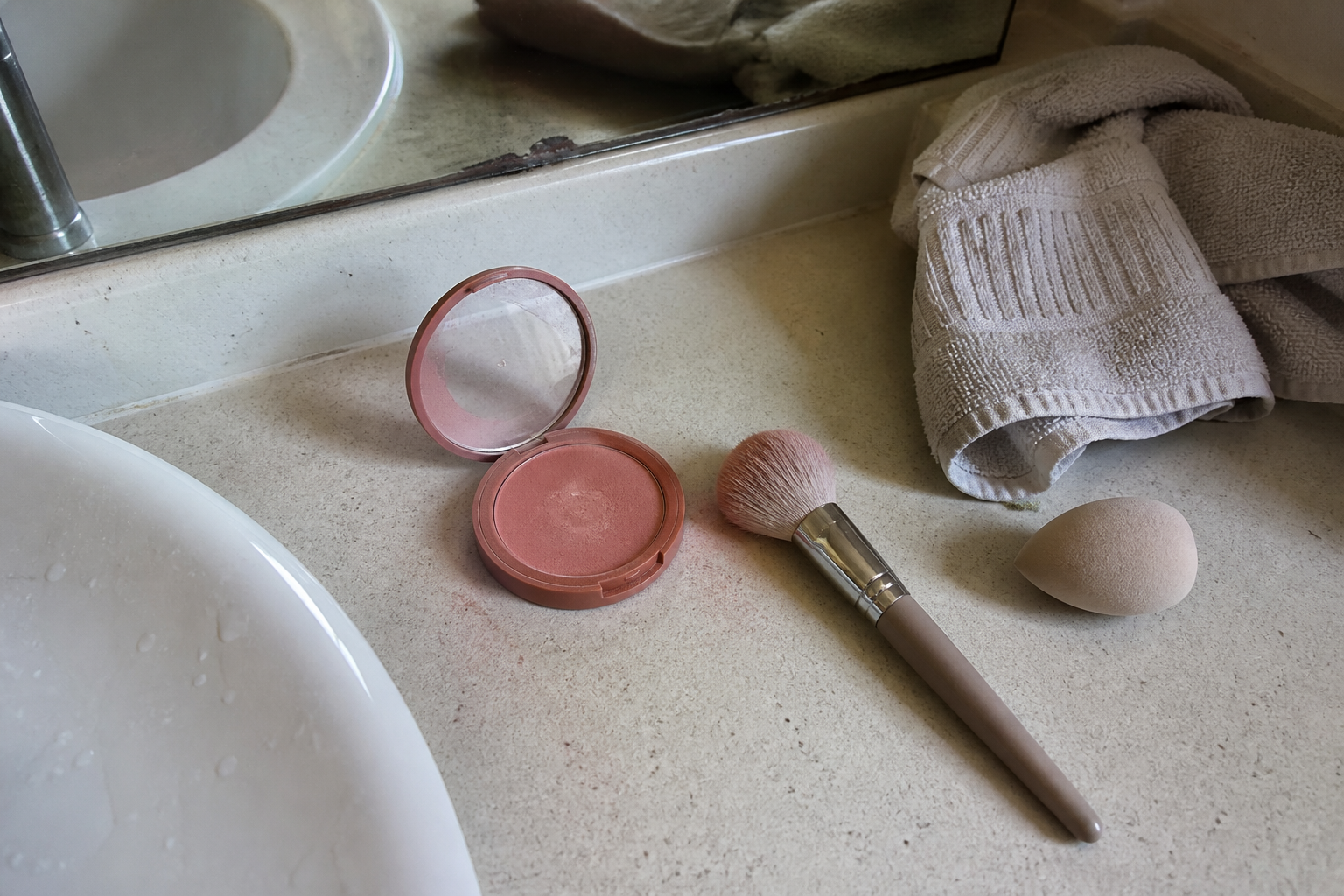 An open rose blush compact, fluffy brush, and makeup sponge on a lived-in bathroom counter.