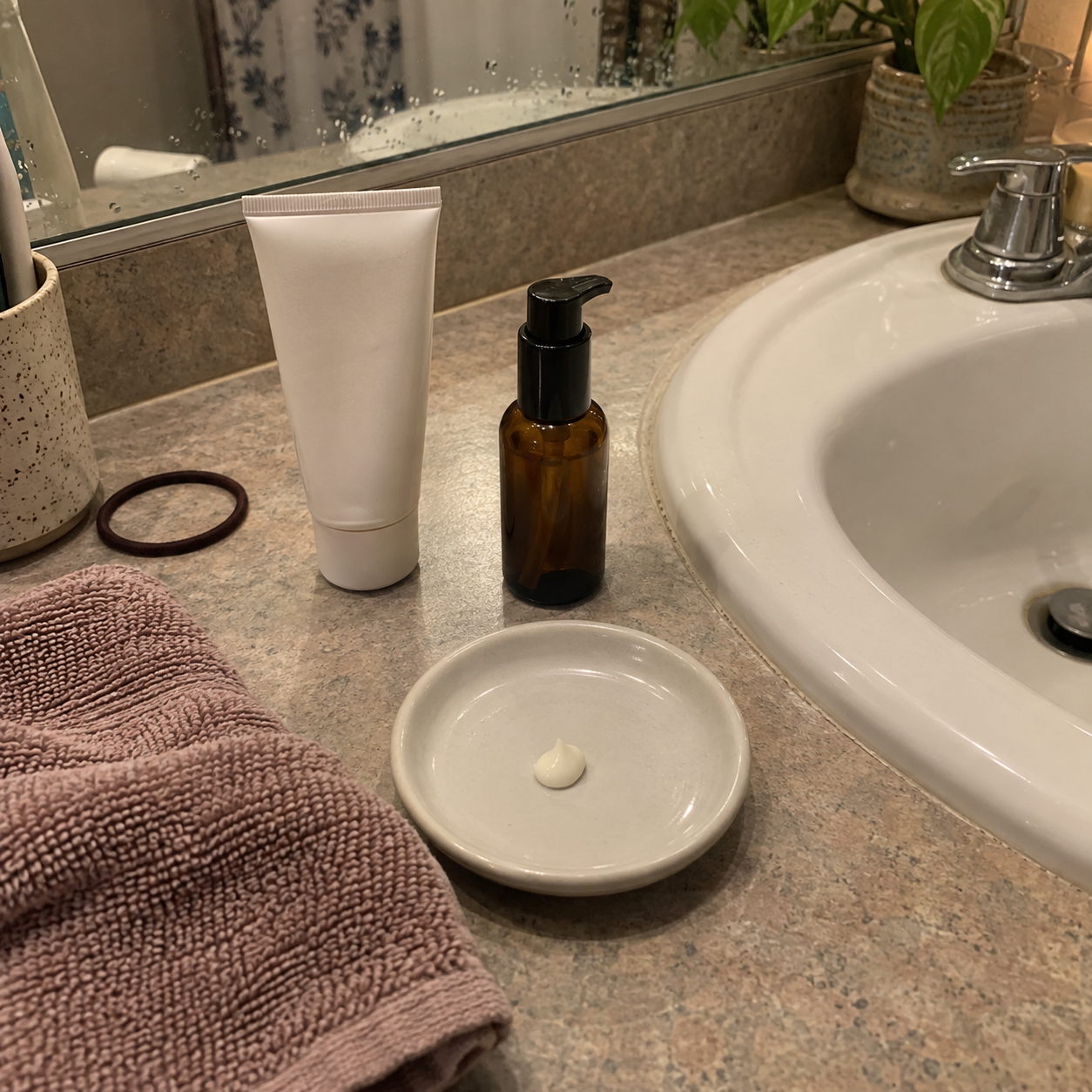 An unbranded moisturizer tube and amber pump bottle beside a small dab of cream on a lived-in bathroom counter.